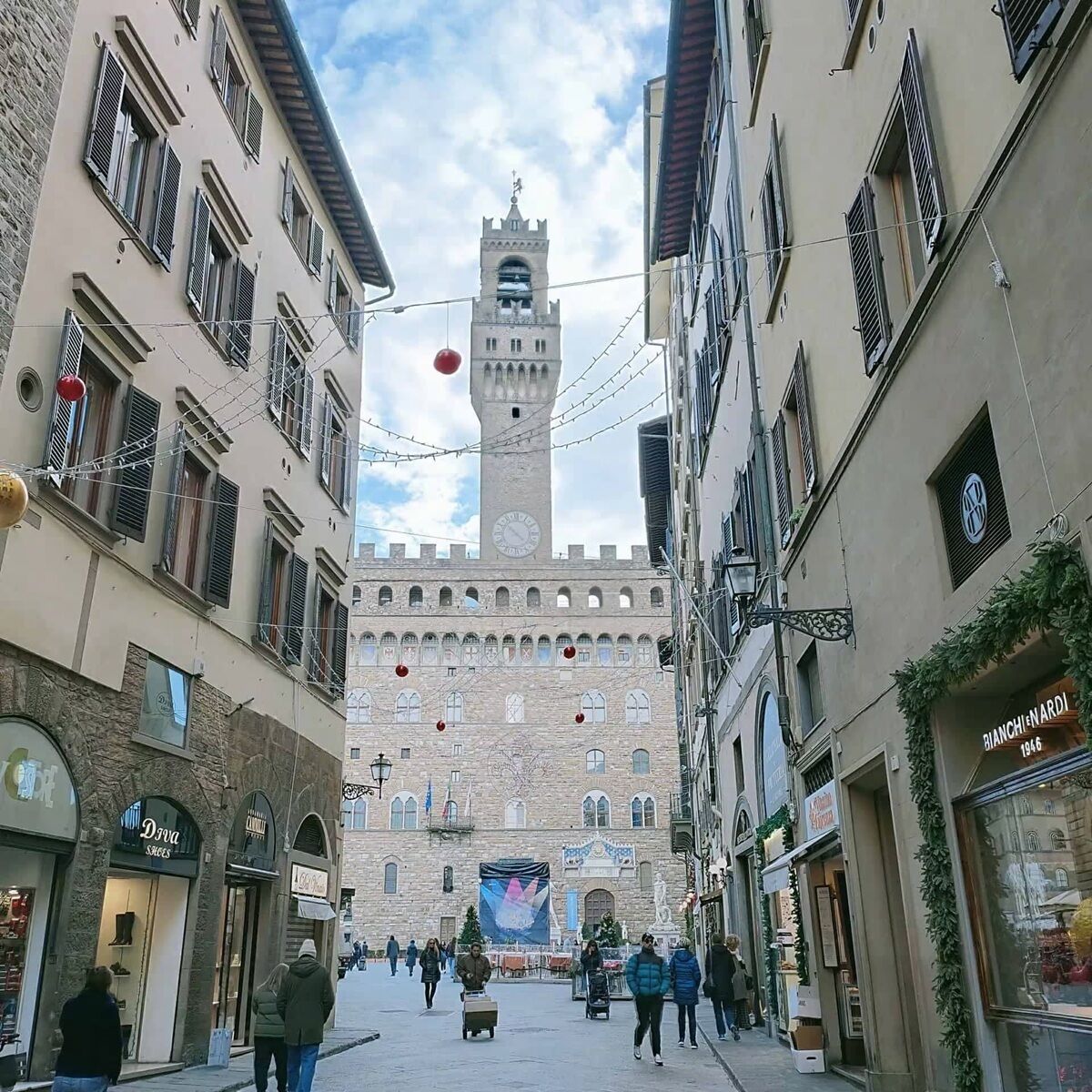 A historic square with a prominent tower, shops on either side, and people walking.