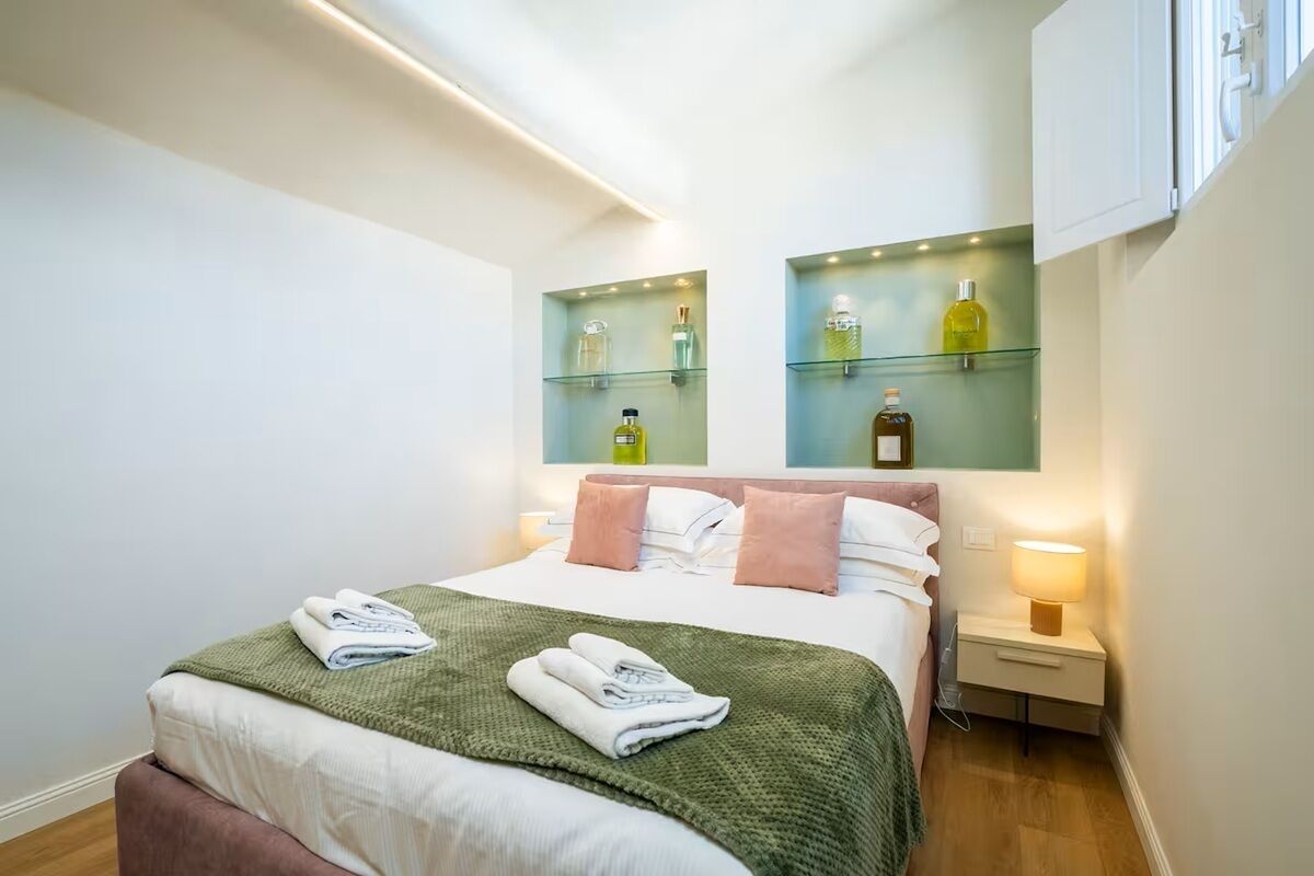 Et hotelværelse med seng, et natbord, en lampe og hylder med toiletartikler.