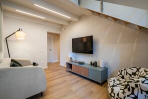 Un salon moderne avec un canapé, un fauteuil à motifs, un téléviseur fixé au mur, une table d’appoint avec une lampe et un plancher en bois.