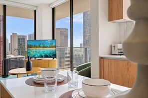 A modern kitchen with a dining table set for two, a television displaying a tropical beach scene, and a view of city buildings through large windows. - The Crosby Worldcenter 2BR Palermo Home  (Miami)