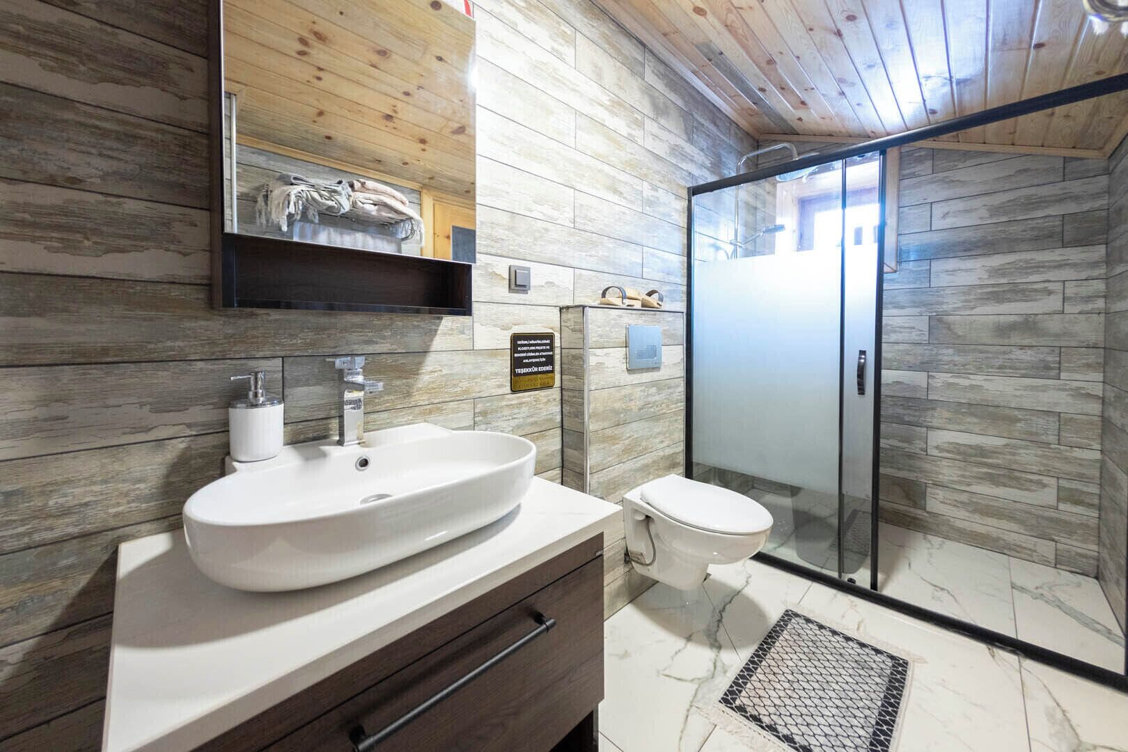 Un baño moderno con pared de madera, un lavamanos blanco, un inodoro y una cabina de ducha con mampara de vidrio.
