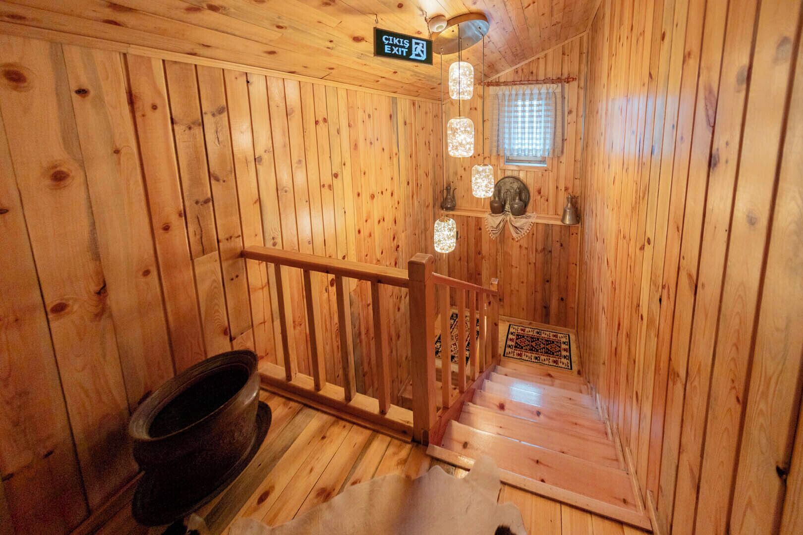 Interior de una cabaña de madera con una escalera de madera, un cartel que indica una salida y una ventana con persiana de cuadros.
