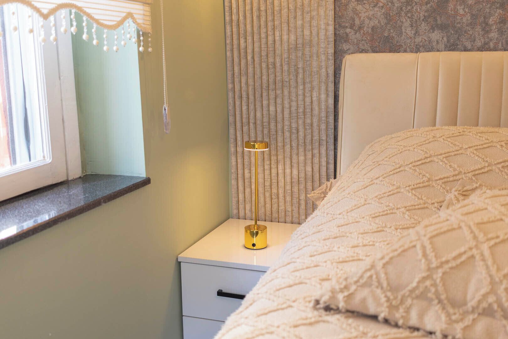 Un dormitorio con cama, una mesita de noche con lámpara, una ventana con cortinas y una pared con textura.