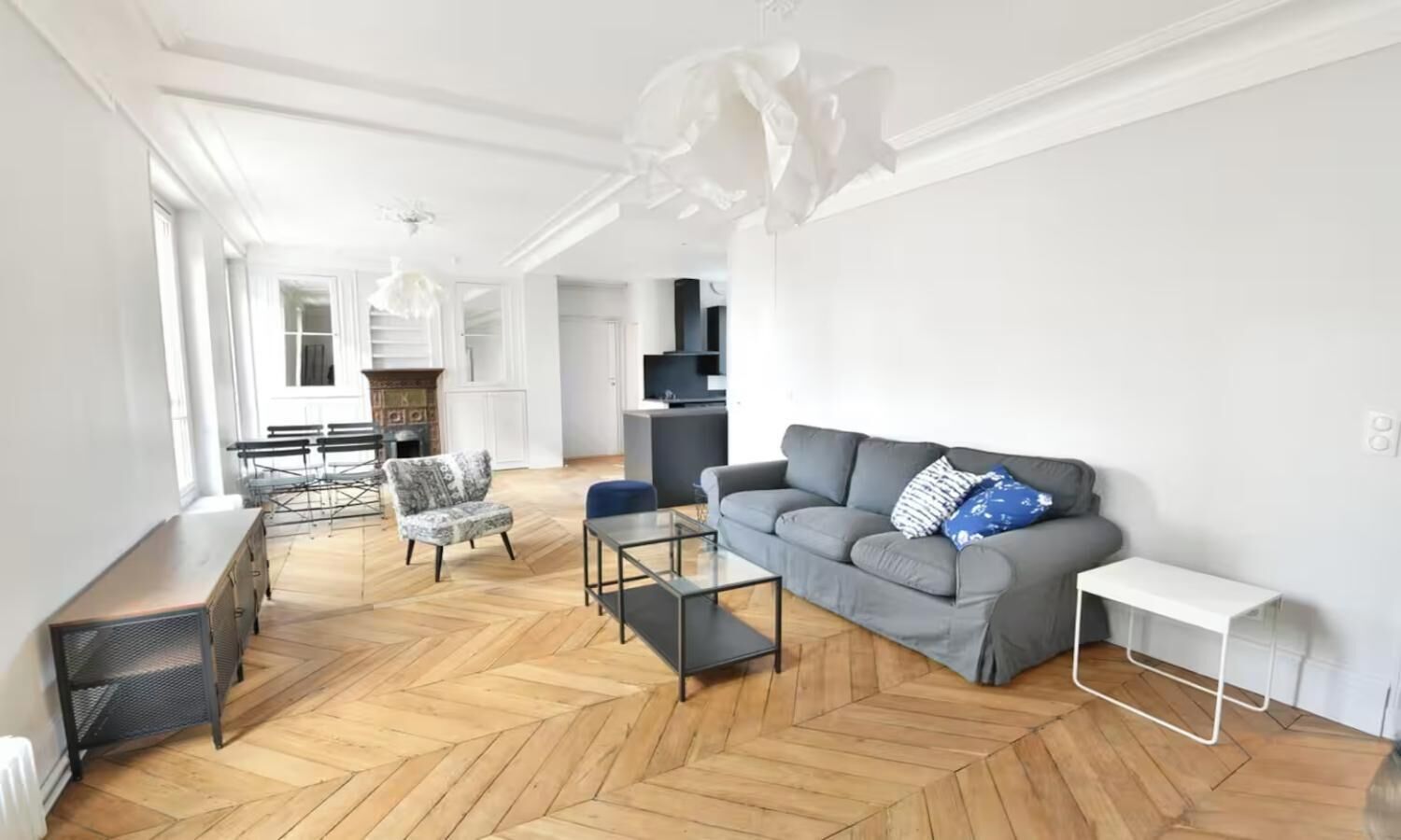 Un salon spacieux doté d’un parquet à motifs en chevrons, d’un canapé gris, d’une table basse et d’un coin repas avec des chaises et une table.