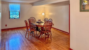 Uma sala de jantar com uma mesa redonda, seis cadeiras de madeira, uma janela com cortinas azuis e um lustre na parede.