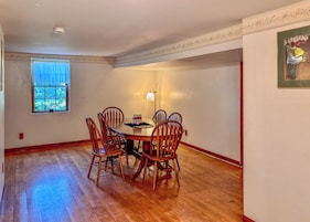 A dining room with a round table, six wooden chairs, a window with blue curtains, and a wall-mounted light fixture.
