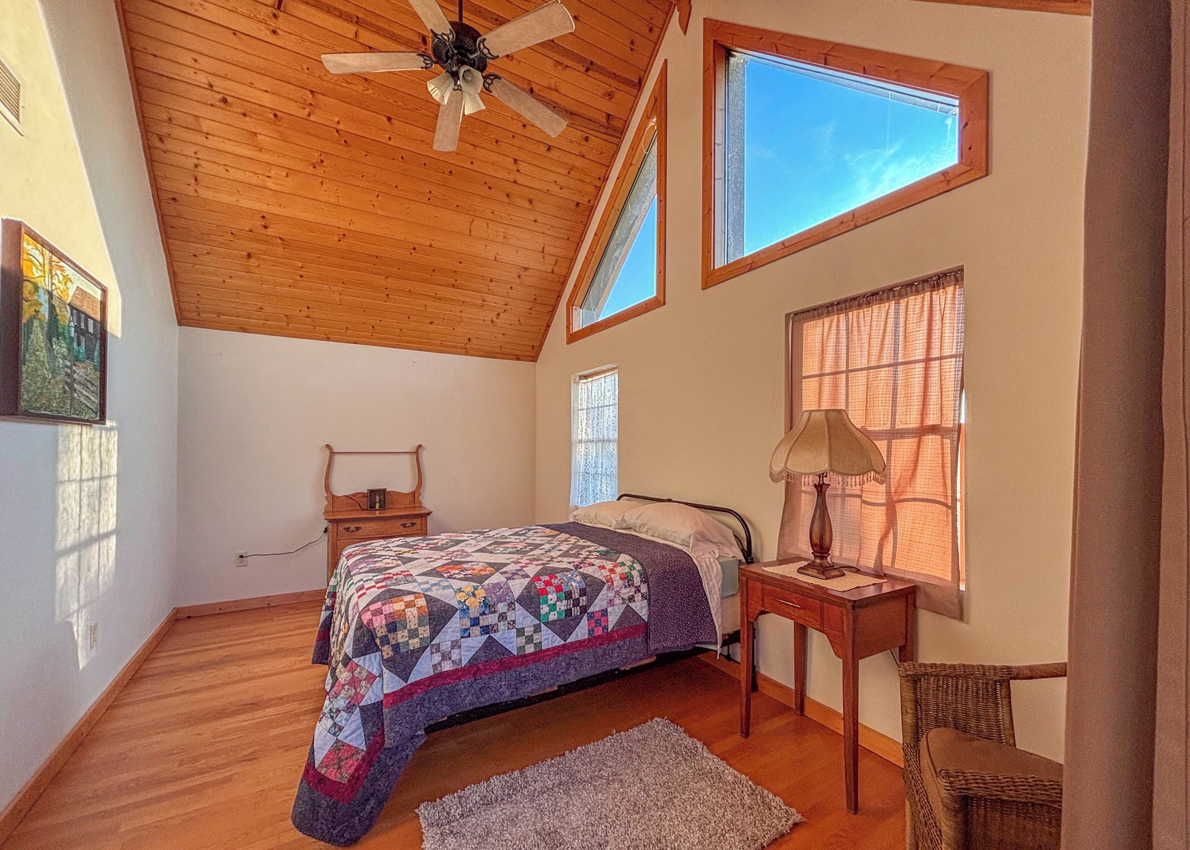 A bedroom with a sloped ceiling, a bed with a quilt, a nightstand, a lamp, a chair, and a window with a view.