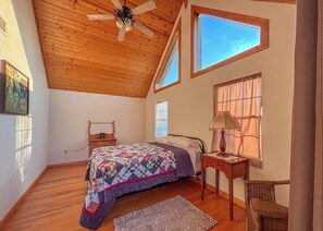 A bedroom with a sloped ceiling, a bed with a quilt, a nightstand, a lamp, a chair, and a window with a view. - Sandberg Retreat (Platteville)