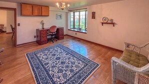 Quarto espaçoso com assoalho de madeira, um tapete azul estampado, uma cadeira de vime, uma escrivaninha com cadeira, uma janela com vista para vegetação e um lustre.