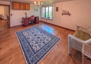 A spacious room with hardwood flooring, a large blue patterned rug, a wicker chair, a desk with a chair, a window with a view of greenery, and a chandelier. - Sandberg Retreat (Platteville)