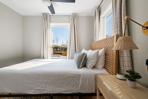 A bedroom with a bed, a bedside table with a lamp, and a view of the outdoors through large windows. - Moultrie & Palmetto Suites | Walk to Bars (Charleston)