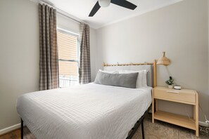 A bedroom with a bed, a nightstand, a lamp, and a window with curtains. - Moultrie & Palmetto Suites | Walk to Bars (Charleston)