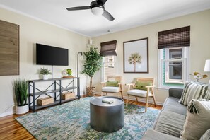 A living room with a flat-screen TV, a ceiling fan, a round coffee table, a sofa, and a large window with blinds. - Moultrie & Palmetto Suites | Walk to Bars (Charleston)