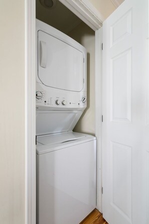 A compact laundry room with a front-loading washer and dryer, a white door, and wooden flooring. - Moultrie & Palmetto Suites | Walk to Bars (Charleston)