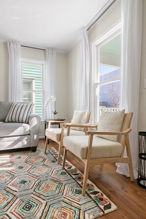 Ein Wohnzimmer mit einer Couch, zwei Sesseln, einem gemusterten Teppich und großen Fenstern mit weißen Vorhängen.