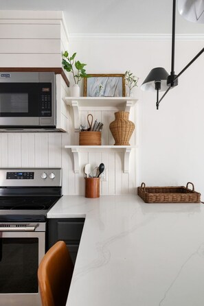 A kitchen with a white countertop, a microwave, an oven, a stove, a hanging light fixture, and shelves with decorative items. - Moultrie & Palmetto Suites | Walk to Bars (Charleston)