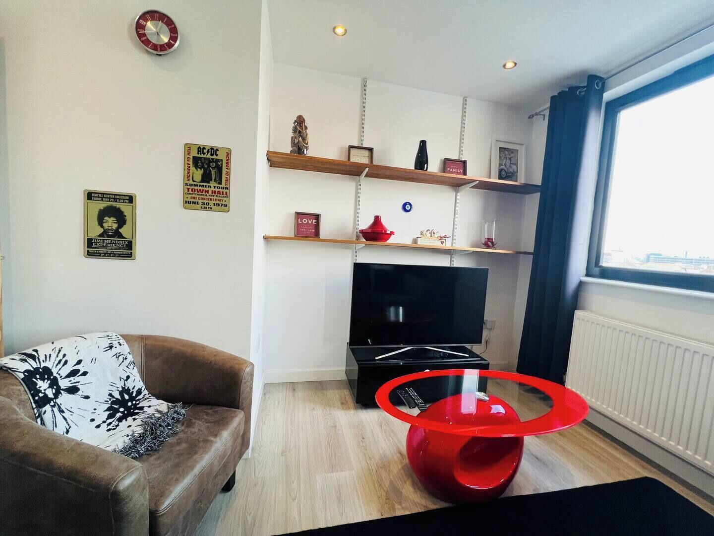 Una sala de estar con un sofá de cuero marrón, un televisor de pantalla plana, un sillón de acento rojo y un ventanal con cortinas.