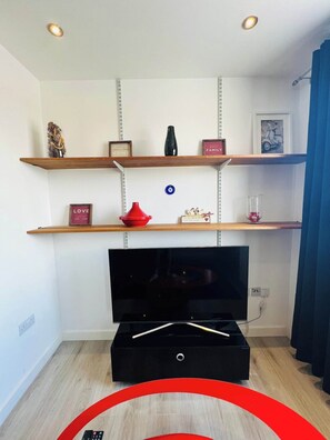 A living room with a flat-screen TV, wooden shelves with decorative items, and a blue curtain. - Modern Flat | Old Town Views, Central Ipswich (Suffolk)