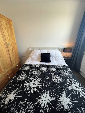 A bedroom with a bed, a wooden wardrobe, a nightstand with a lamp, and a window with curtains. - Modern Flat | Old Town Views, Central Ipswich (Suffolk)