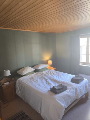 Een slaapkamer met een houten plafond, een bed met wit beddengoed, een nachtkastje met een lamp en een raam met gordijnen.