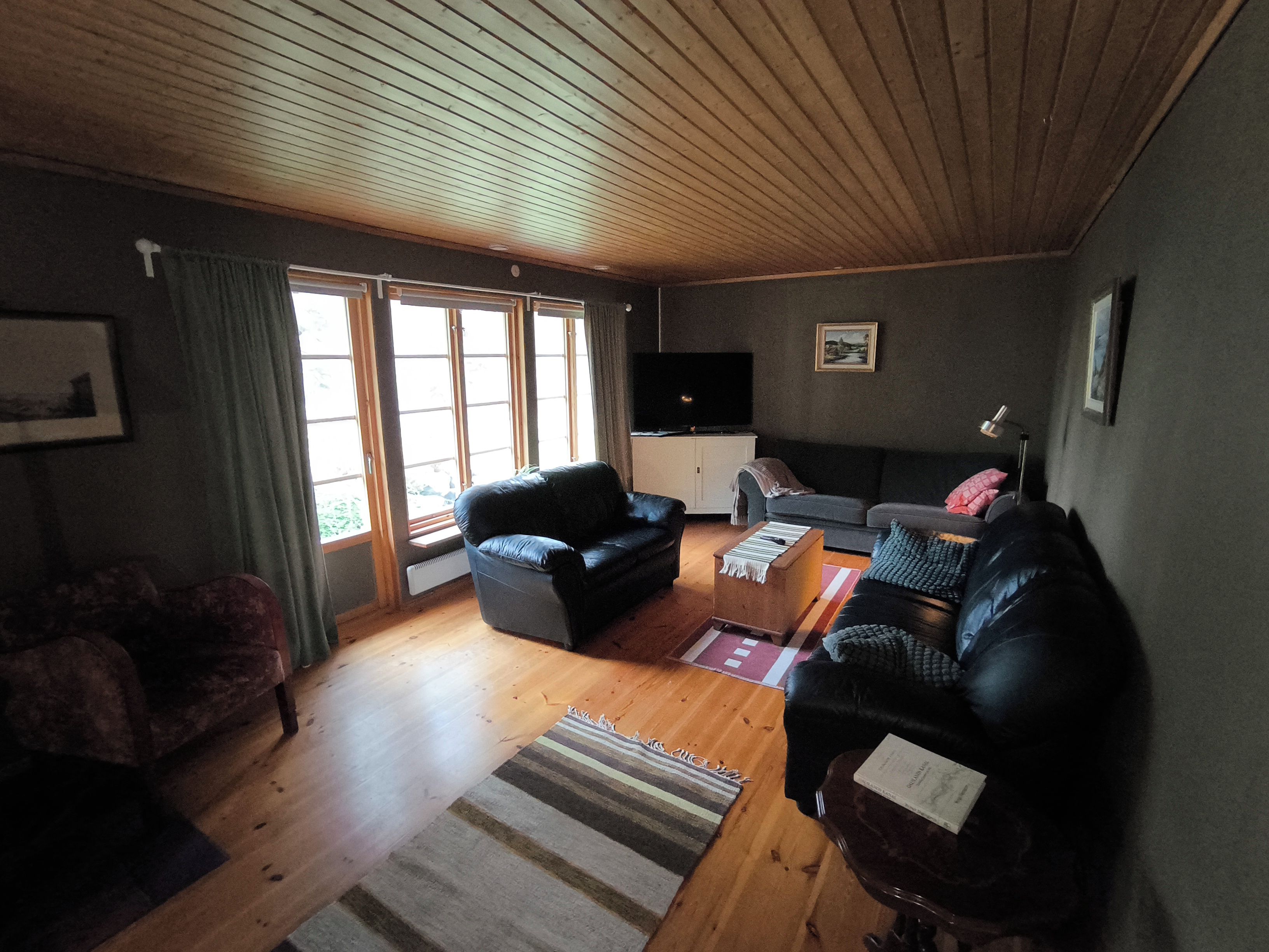 Un salon avec un plafond en bois, un canapé en cuir noir, un canapé gris, une petite table et un téléviseur posé sur un meuble.