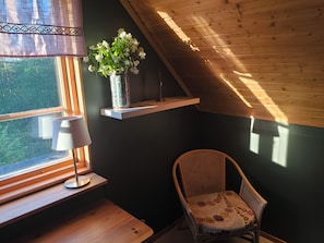 Une pièce avec un plafond en pente, une fenêtre donnant sur un paysage verdoyant, une chaise avec un coussin à motifs et un vase rempli de fleurs posé sur une étagère.