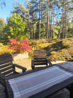 Terraza de madera con dos sillas y una mesa, con vista a un área boscosa que incluye un muro de piedra y diversa vegetación.