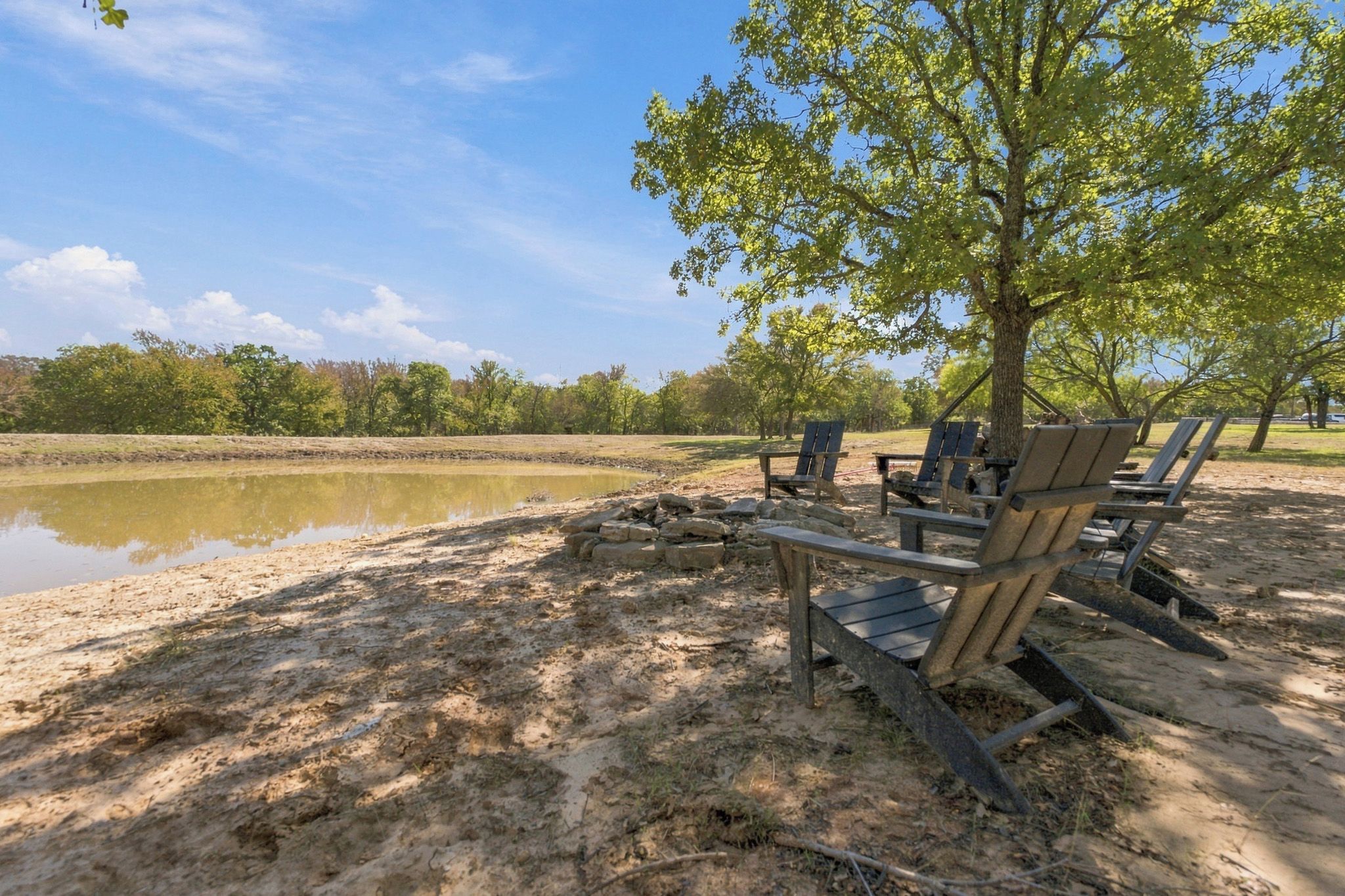 Un paisaje junto al lago con sillas de madera y una mesa de picnic, rodeado de árboles y un cielo despejado.