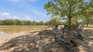 Un paisaje junto al lago con sillas de madera y una mesa de picnic, rodeado de árboles y un cielo despejado.