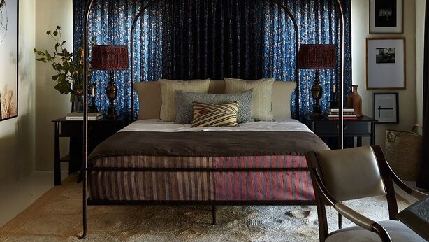 Ein Schlafzimmer mit Himmelbett, großem Fenster mit Vorhängen, Nachttisch mit Lampe und gerahmten Bildern an der Wand.