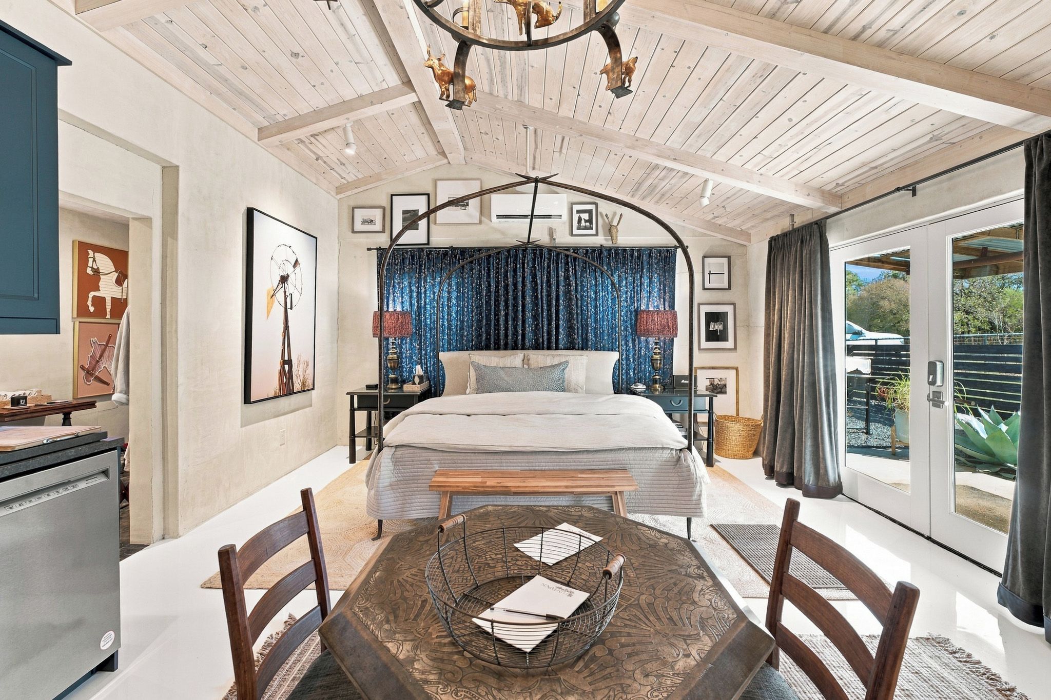 A bedroom with a bed, a dining table with chairs, a chandelier, and a view of the outside.