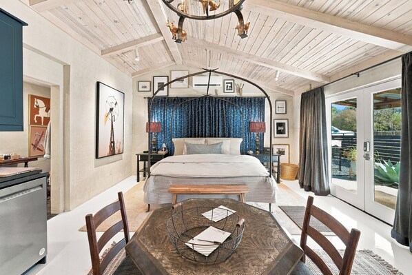 A bedroom with a bed, a dining table with chairs, a chandelier, and a view of the outside.