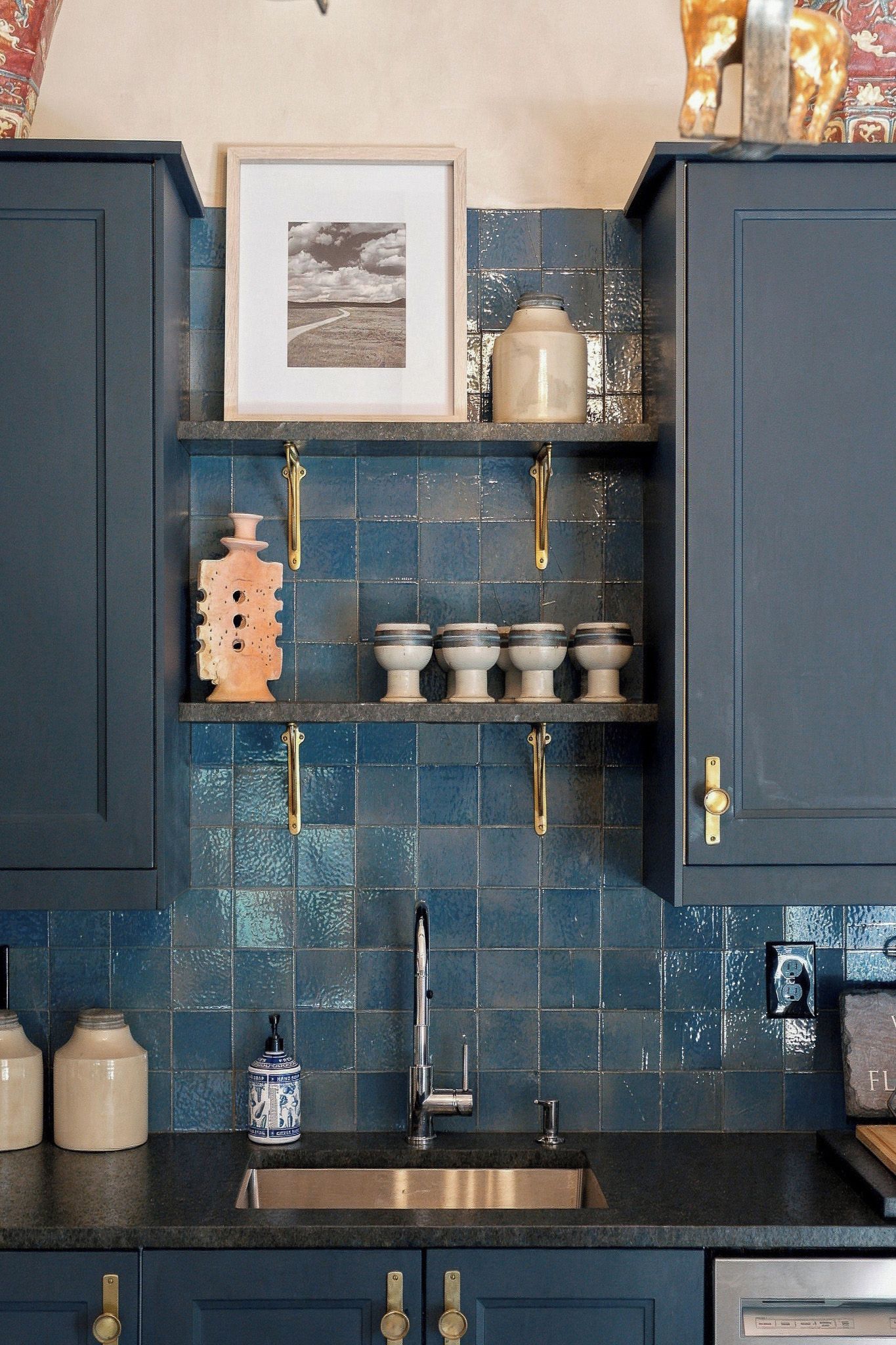 A kitchen with dark blue cabinets, a tiled backsplash, a stainless steel sink, and various kitchen items on shelves.