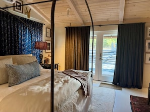 A bedroom with a bed, a nightstand, a lamp, and a view of the outdoors through a door.
