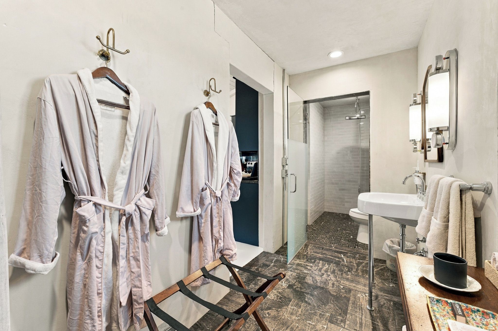 A hotel bathroom with two robes hanging on hooks, a shower area, a sink with a mirror, and a wooden floor.