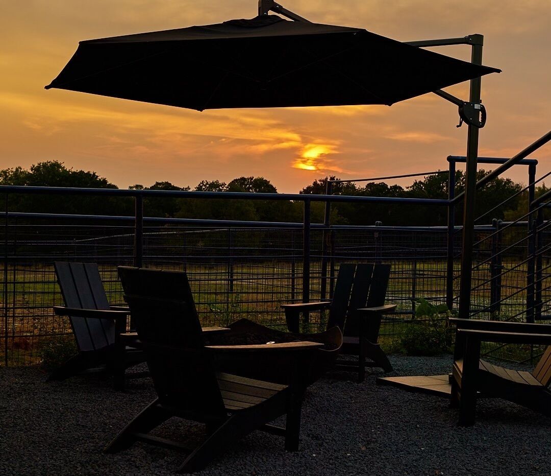 An outdoor seating area with chairs and an umbrella, overlooking a fenced field with a sunset view.