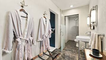 A hotel bathroom with two robes hanging on hooks, a shower area, a sink with a mirror, and a wooden floor.