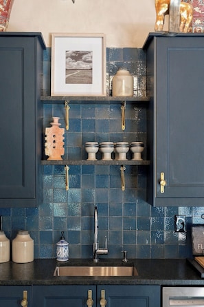 A kitchen with dark blue cabinets, a tiled backsplash, a stainless steel sink, and various kitchen items on shelves.