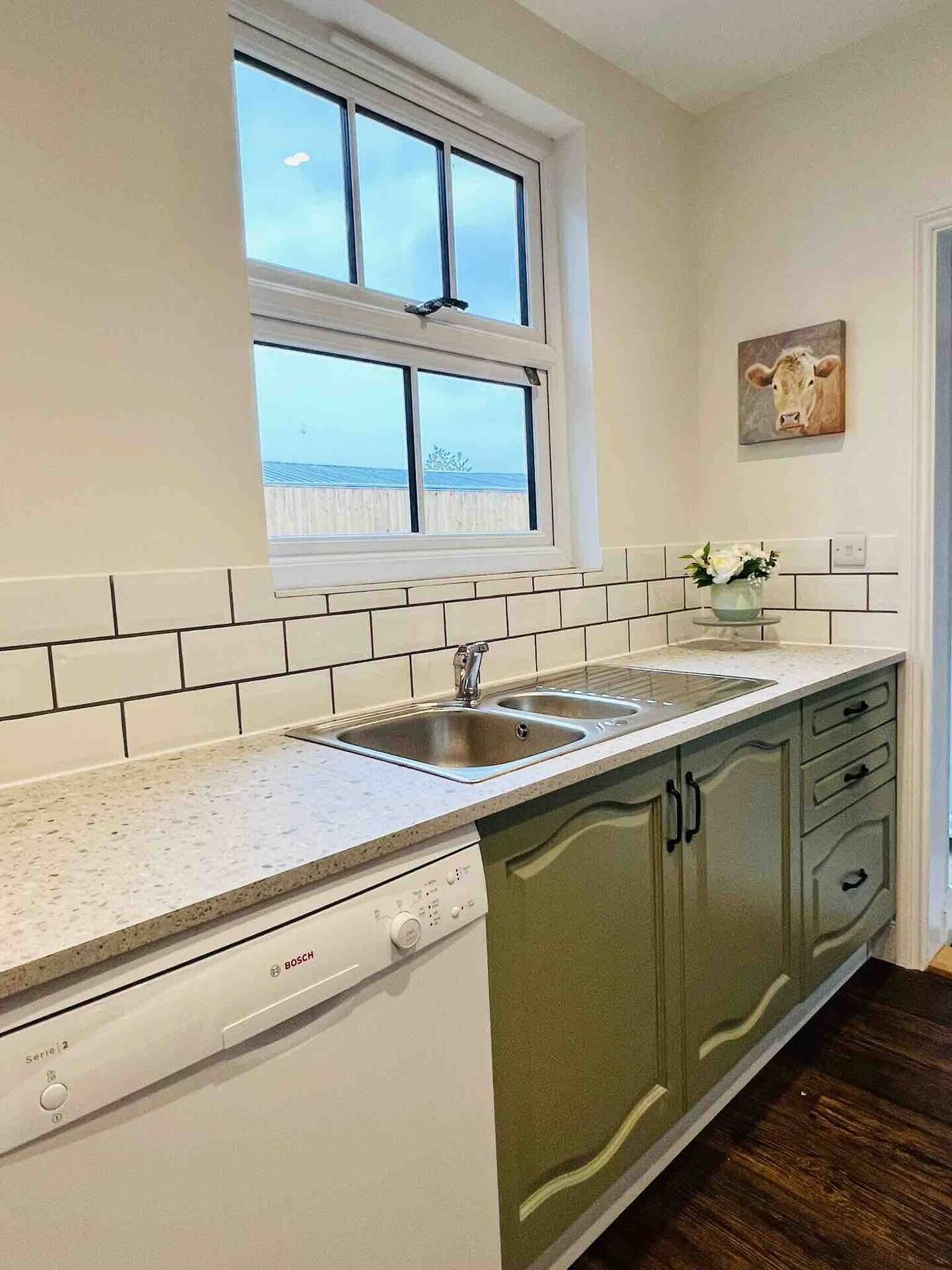 A kitchen with a Bosch dishwasher, a window with a view, and a cow painting on the wall.