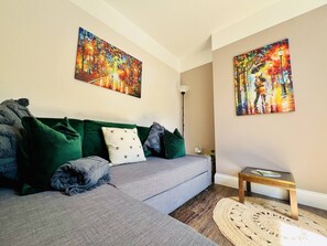 Uma sala de estar com um sofá cinza, almofadas verdes e cinzas, um tapete redondo tecido e duas pinturas nas paredes.