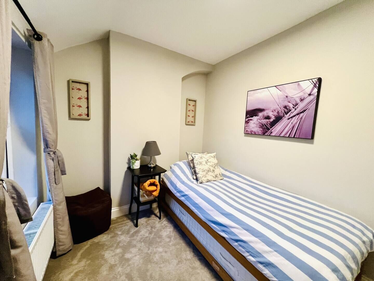 A bedroom with a single bed, a nightstand with a lamp, a framed picture on the wall, and a window with curtains.