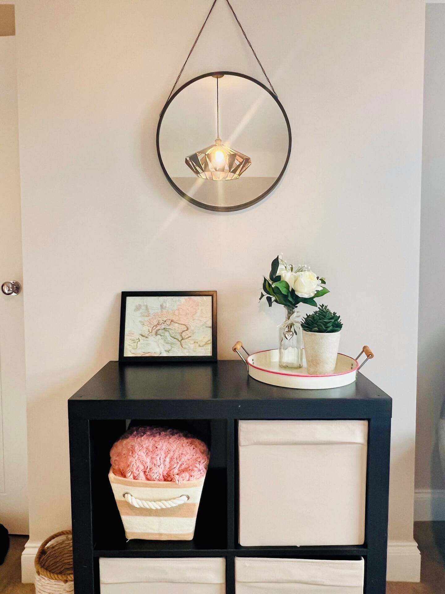 Et sort konsolbord med en hængende lampe over, et indrammet billede, en vase med hvide blomster og en kurv med lyserøde ting.