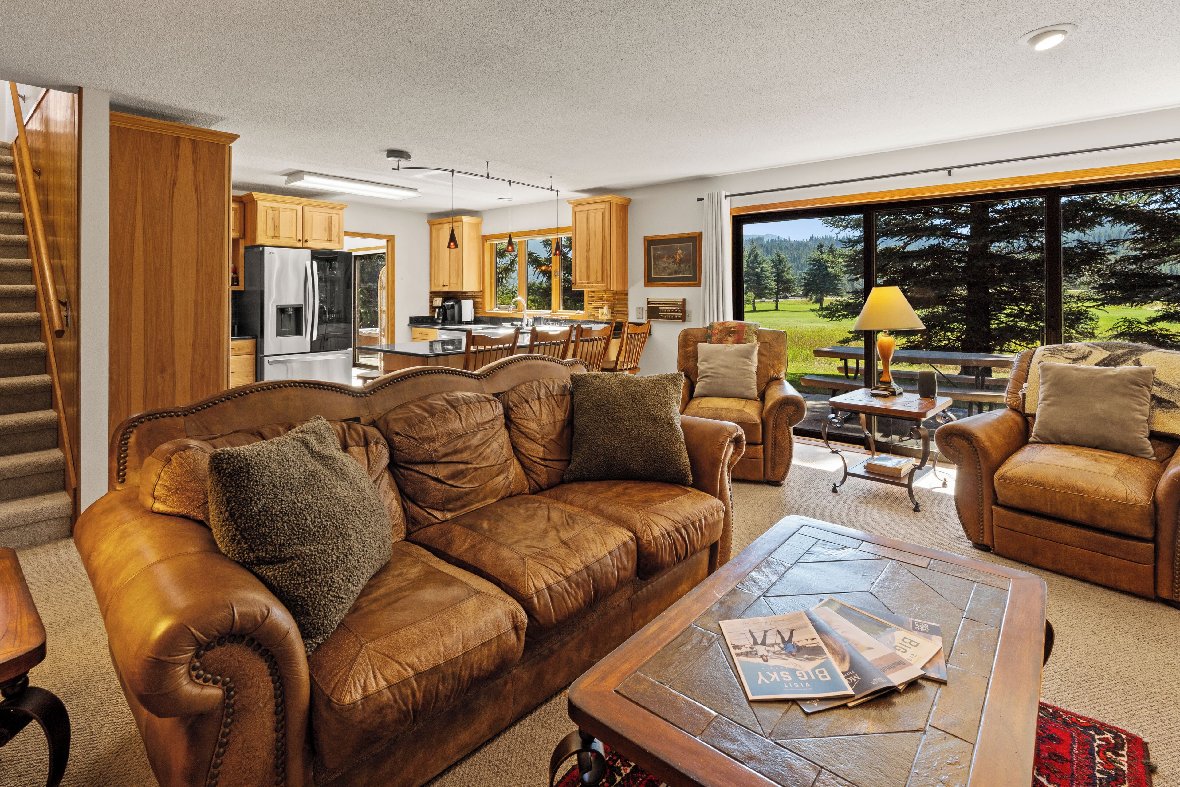 A spacious living room with a brown leather sofa, a coffee table with magazines, and a view of a kitchen and outdoor area through large windows.