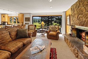 A spacious living room with a fireplace, leather sofas, a coffee table with magazines, and a view of a forest through large windows. - Charming 5-bedroom house in wonderful Big Sky for your getaway (Big Sky)