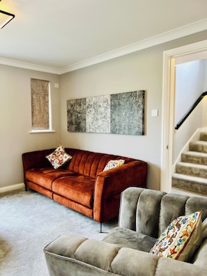 A living room with a brown sofa and a grey armchair, both with patterned cushions. A large abstract painting hangs on the wall above the sofa. - Boutique Home & Private Garden | City Escape (Colchester)