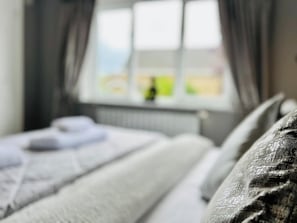 A bed with pillows and a blanket, a window with curtains, and a view of a landscape. - Boutique Home & Private Garden | City Escape (Colchester)