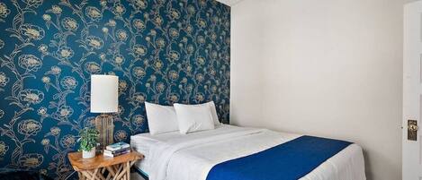A bedroom with a bed, bedside table, lamp, and a blue floral wallpaper.