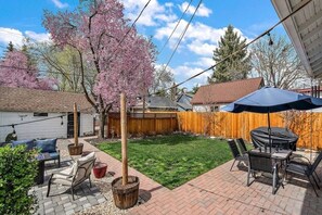Un cortile con un'area patio, una recinzione in legno, un albero con fiori rosa e un angolo barbecue.