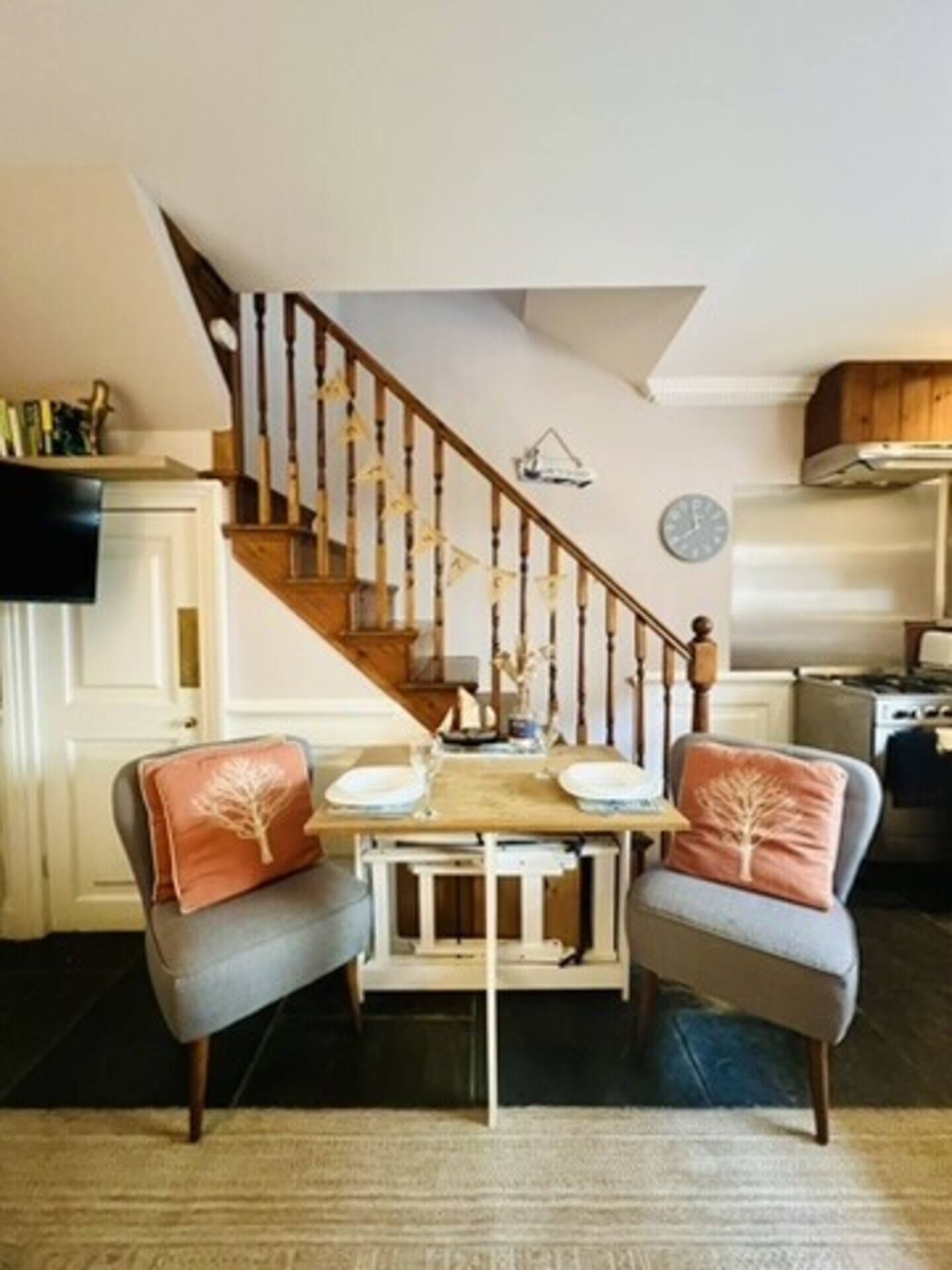 A dining area with a wooden staircase, a clock on the wall, and a kitchen in the background.
