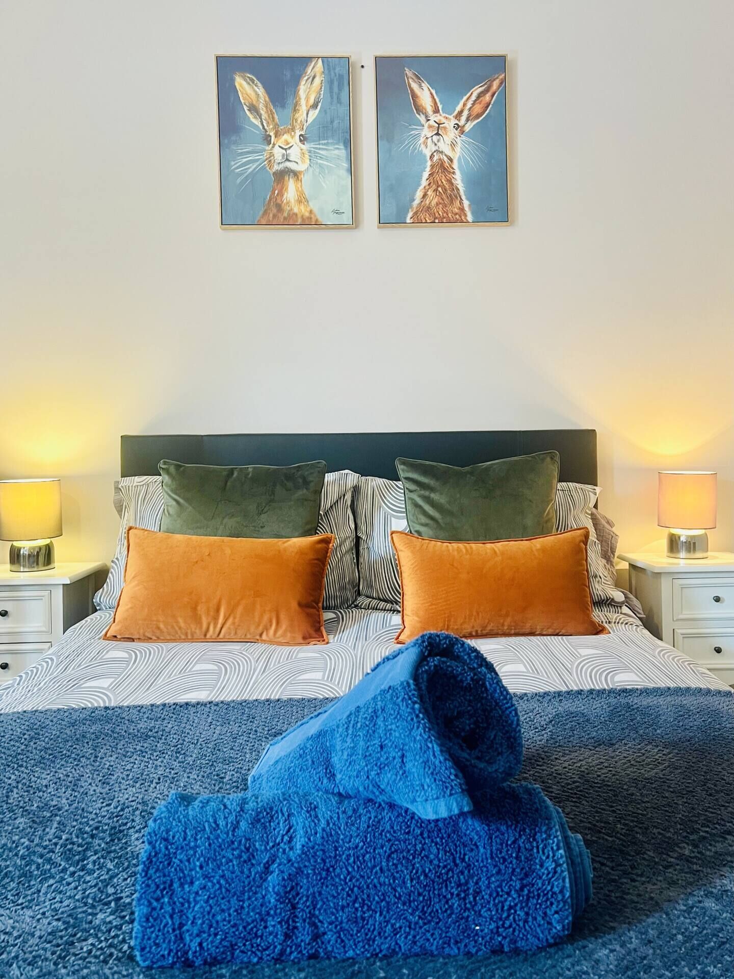Un lit avec une literie à motifs, deux oreillers, une couverture bleue et deux tableaux représentant des lapins, accrochés au mur.